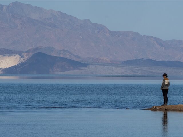 A person standing beside still blue water beneath a vast range of desert mountains